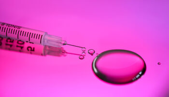 Syringe and neurotoxin droplet on a rich pink background, representing preventive Botox, wellness, & skincare trends