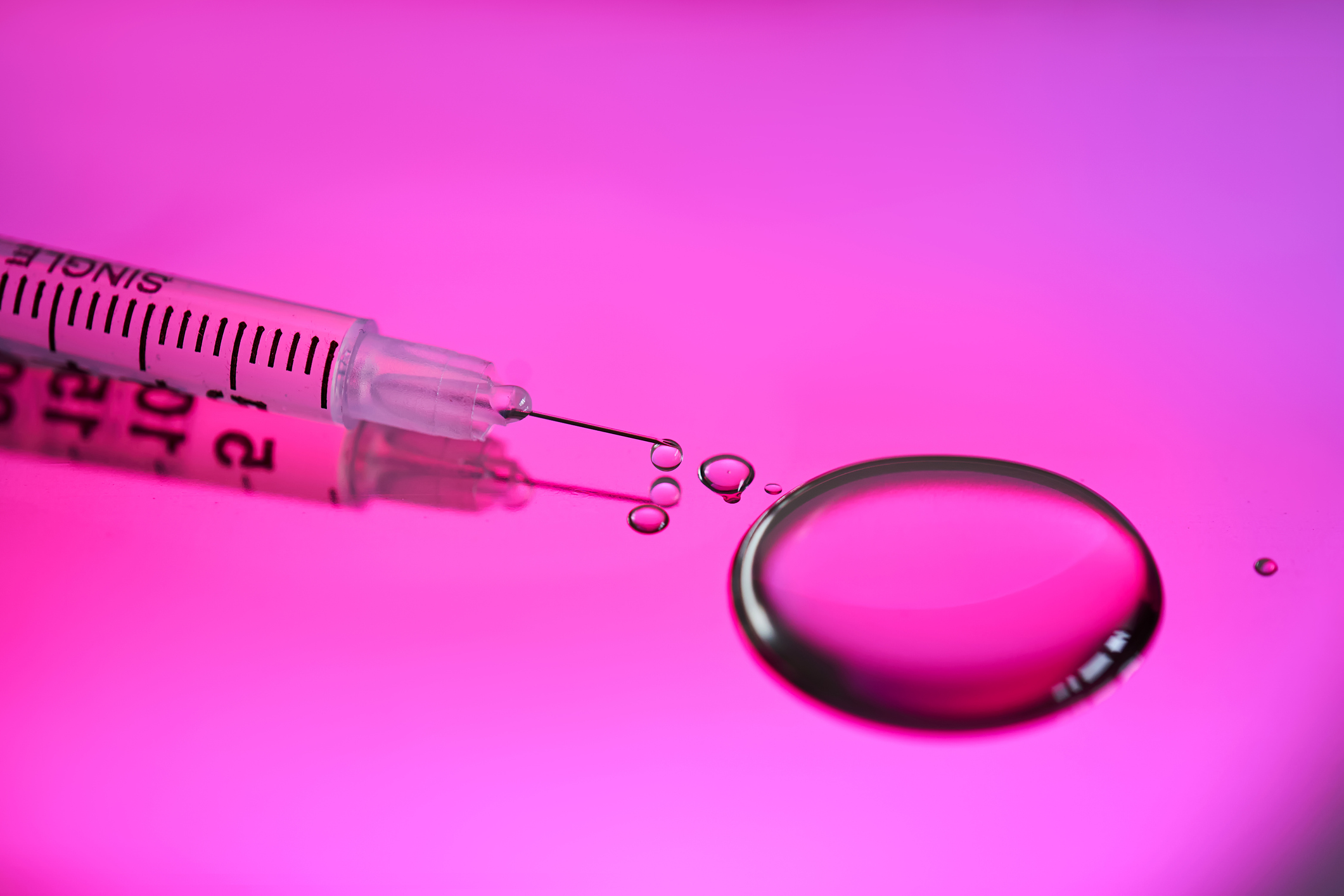 Syringe and neurotoxin droplet on a rich pink background, representing preventive Botox, wellness, & skincare trends